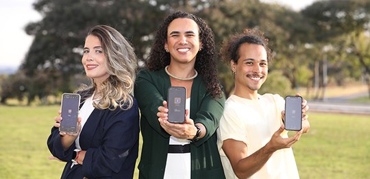 A imagem mostra três pessoas em um ambiente externo, lado a lado, sorrindo e segurando celulares...